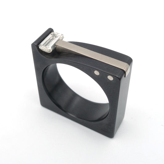 Strakke blokvormige ring van zwart zirkonium met een witgouden inlay, gezet met een emerald geslepen diamant op een witte achtergrond.