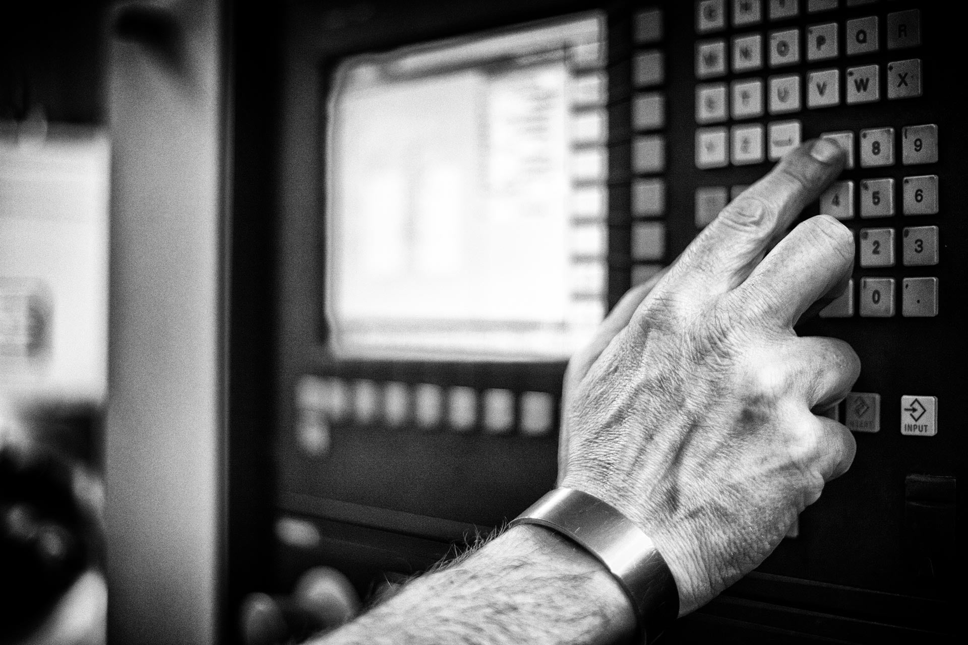Joeri Dijkman bedient een CNC-machine door op het bedieningspaneel te drukken – zwart-witfoto van het productieproces.

